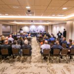 A conference room with people seated facing a panel of speakers at the front. The panelists are at a long table with microphones, and a person is speaking at a podium. The audience appears attentive, with several rows of chairs filled.