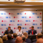 Five people seated at a table in a conference setting with microphones, participating in a panel discussion. Behind them is a banner with logos, and an audience is visible in the foreground.
