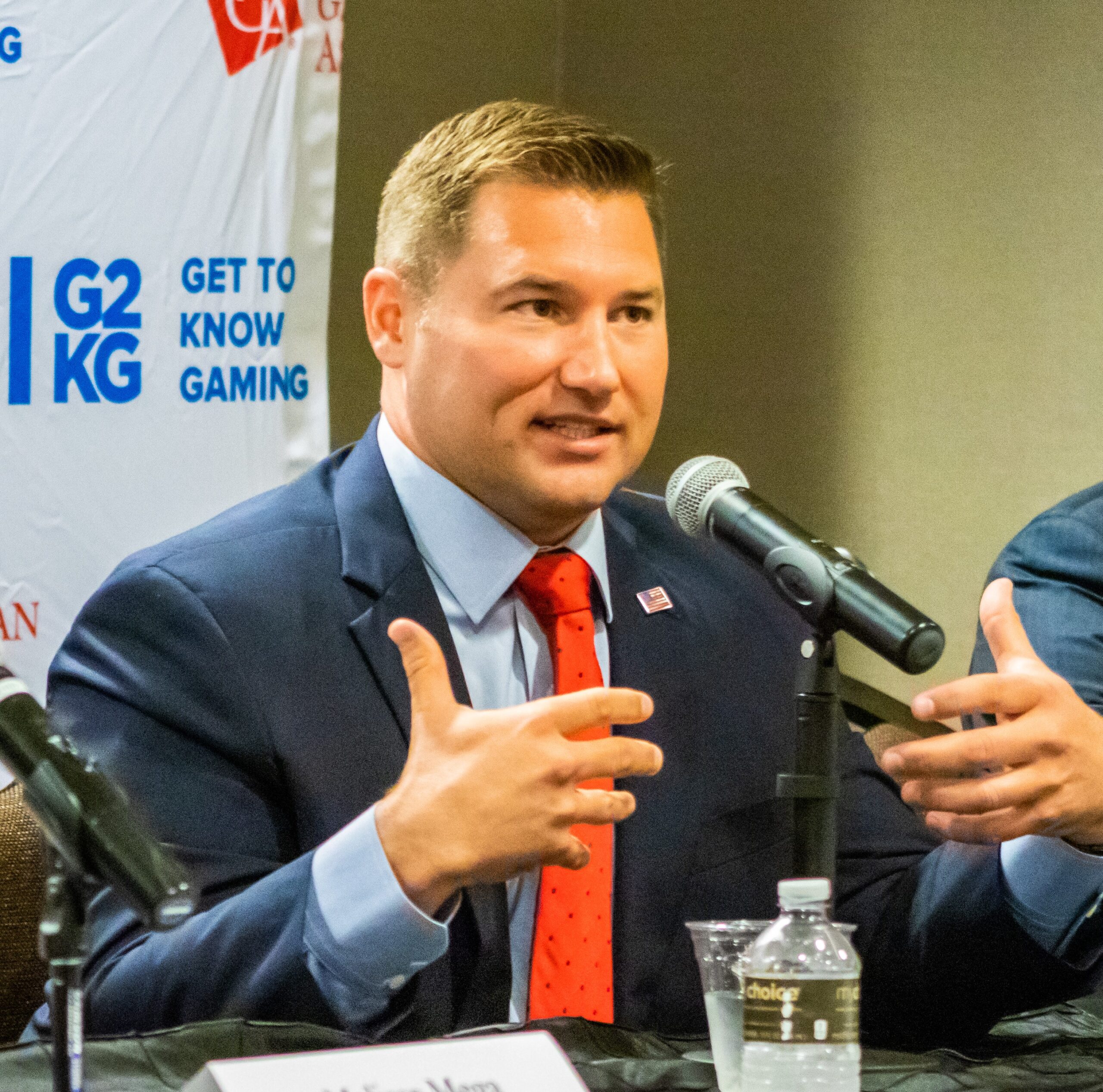 A man in a suit and red tie speaks into a microphone at a panel. He gestures with his hands while sitting at a table with a water bottle. A logo and text about gaming are on the backdrop.
