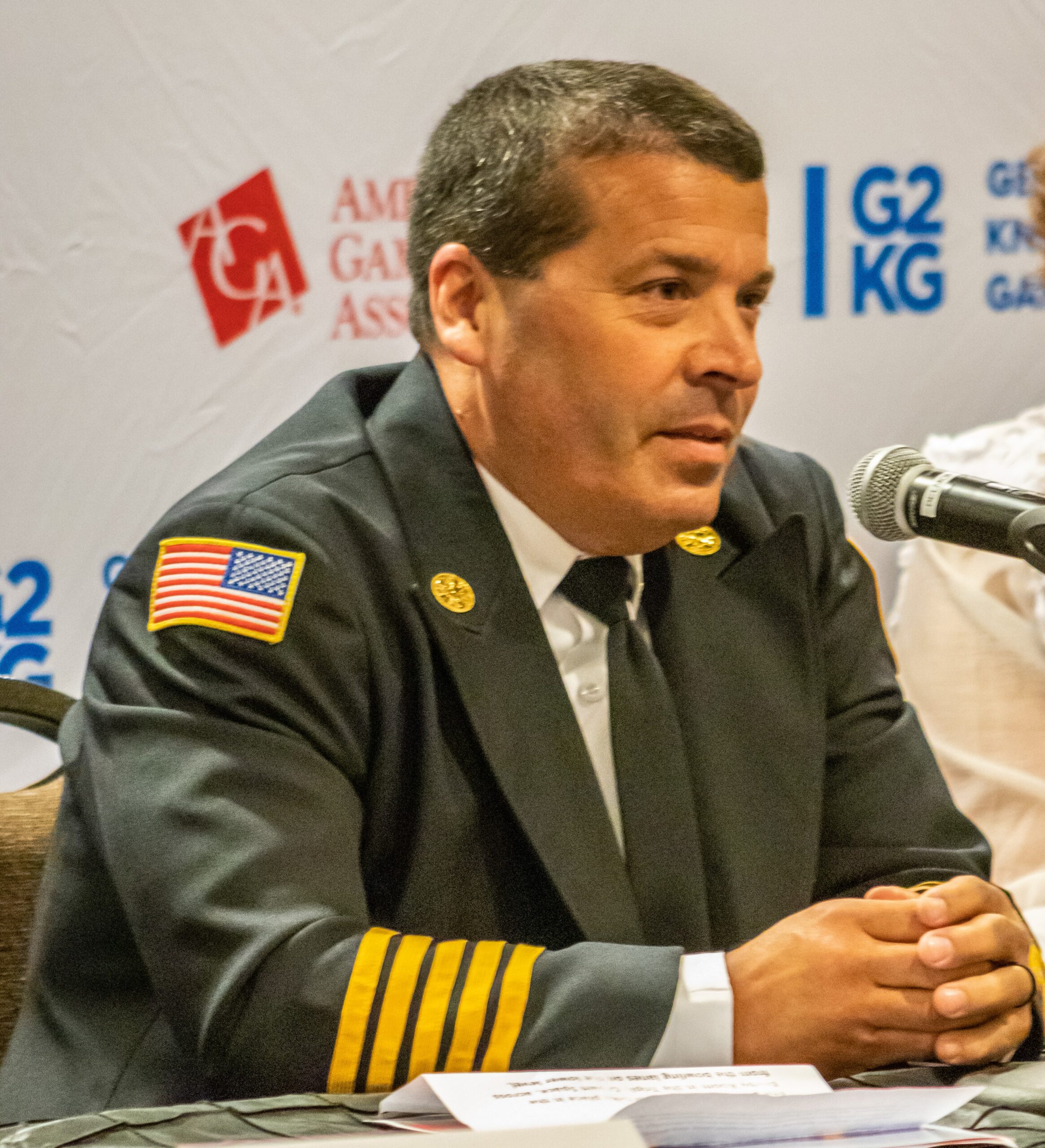 A man in a firefighter uniform with an American flag patch on the sleeve is speaking into a microphone at a press conference or event. He is seated at a table with a backdrop displaying the G2E and other logos.