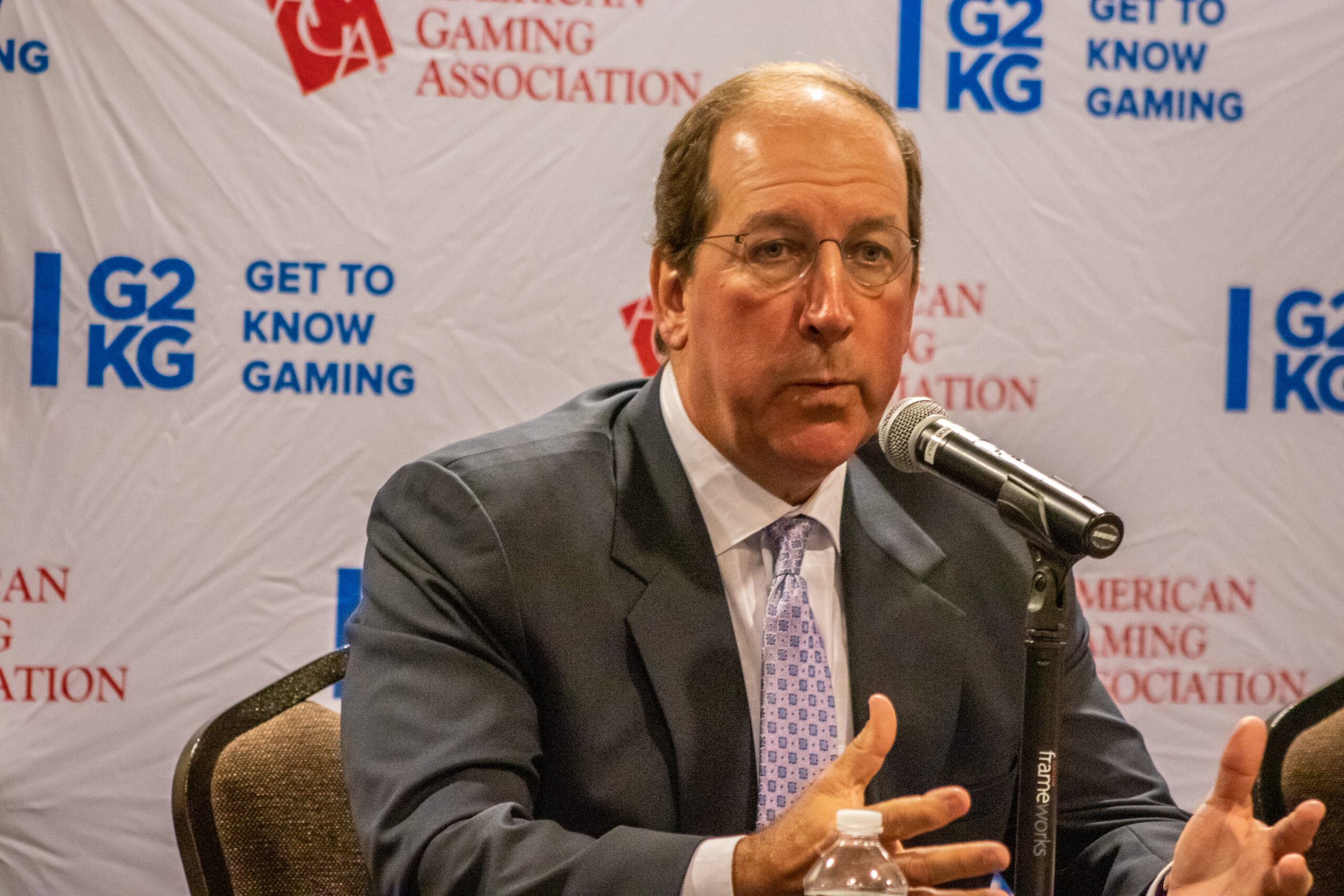 A man in a suit speaks into a microphone at a press conference. The background features logos for 'American Gaming Association' and 'Get to Know Gaming.' He gestures with his hands while addressing the audience.