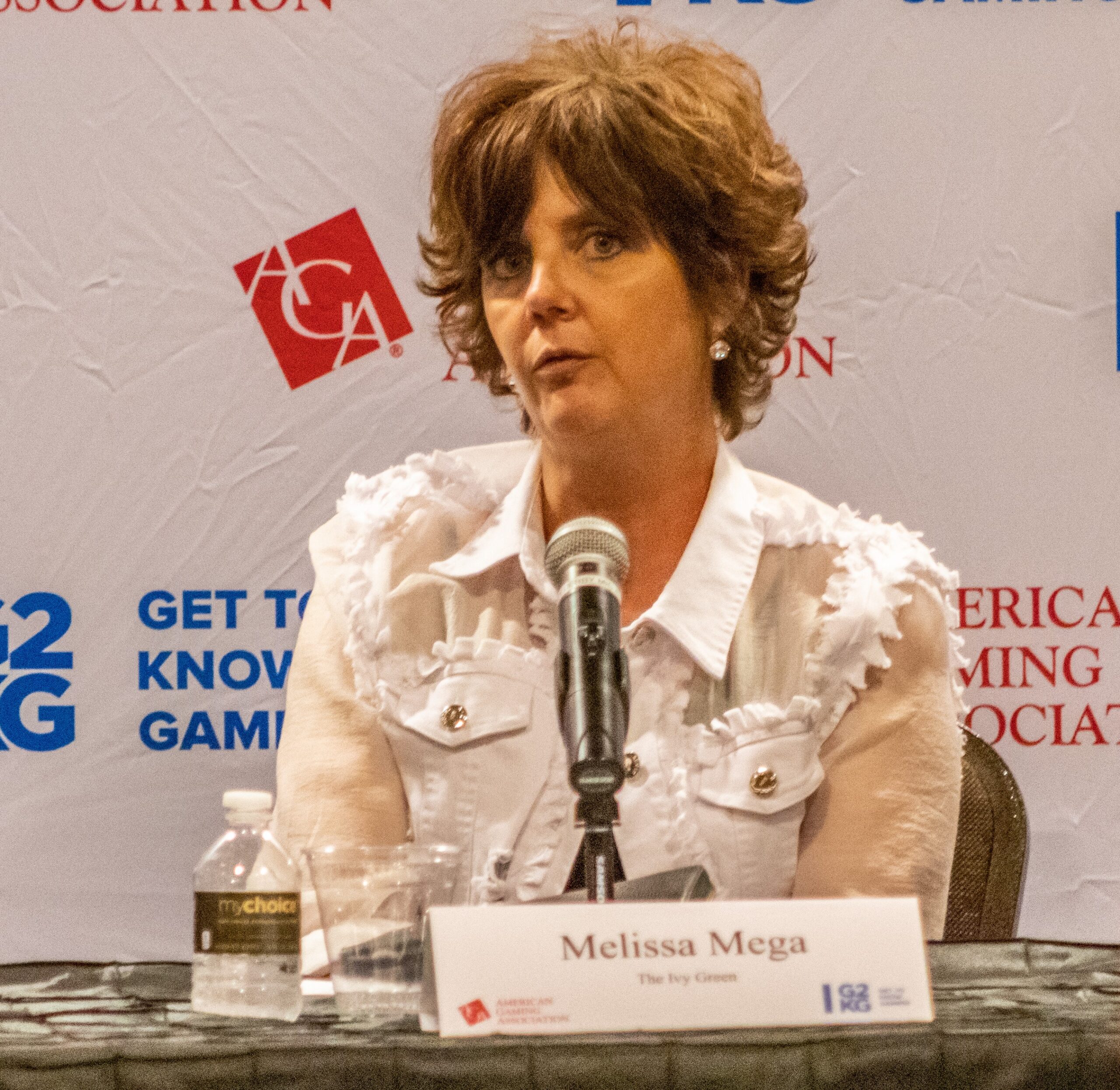 A woman with short brown hair sits at a table with a microphone in front of her. Behind her is a backdrop with various logos and text. A name card on the table reads "Melissa Mega." There is a bottle of water and a glass next to her.