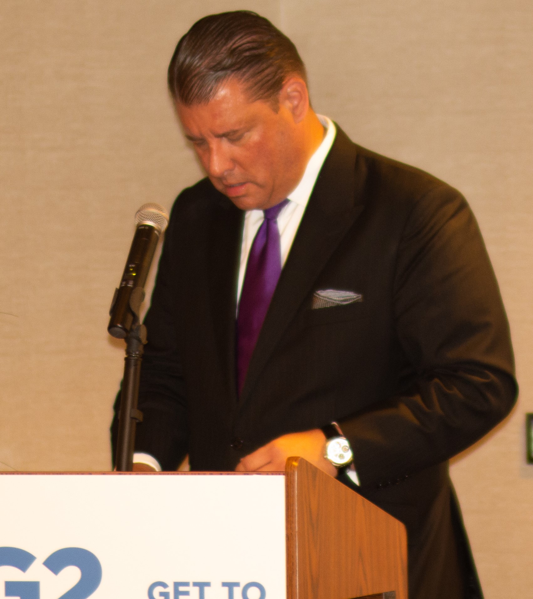 A man in a black suit and purple tie stands at a podium, speaking into a microphone. He has slicked-back hair and is appearing to read from notes or documents on the podium. A partial view of a sign with blue text is visible in front.