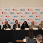 A panel of seven individuals seated behind a long table, engaged in a discussion. They are in a conference room with a backdrop featuring logos of the American Gaming Association. Audience members are visible in the foreground.