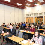 A group of people sitting at rectangular tables in a conference room with white walls and a red ceiling. Some attendees are taking notes, while others are using electronic devices or watching the front of the room.