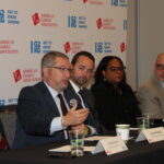 A group of six people seated at a panel discussion with microphones. They are in front of a backdrop featuring the American Gaming Association logo. One man is speaking, gesturing with his hand. Water bottles and nameplates are on the table.
