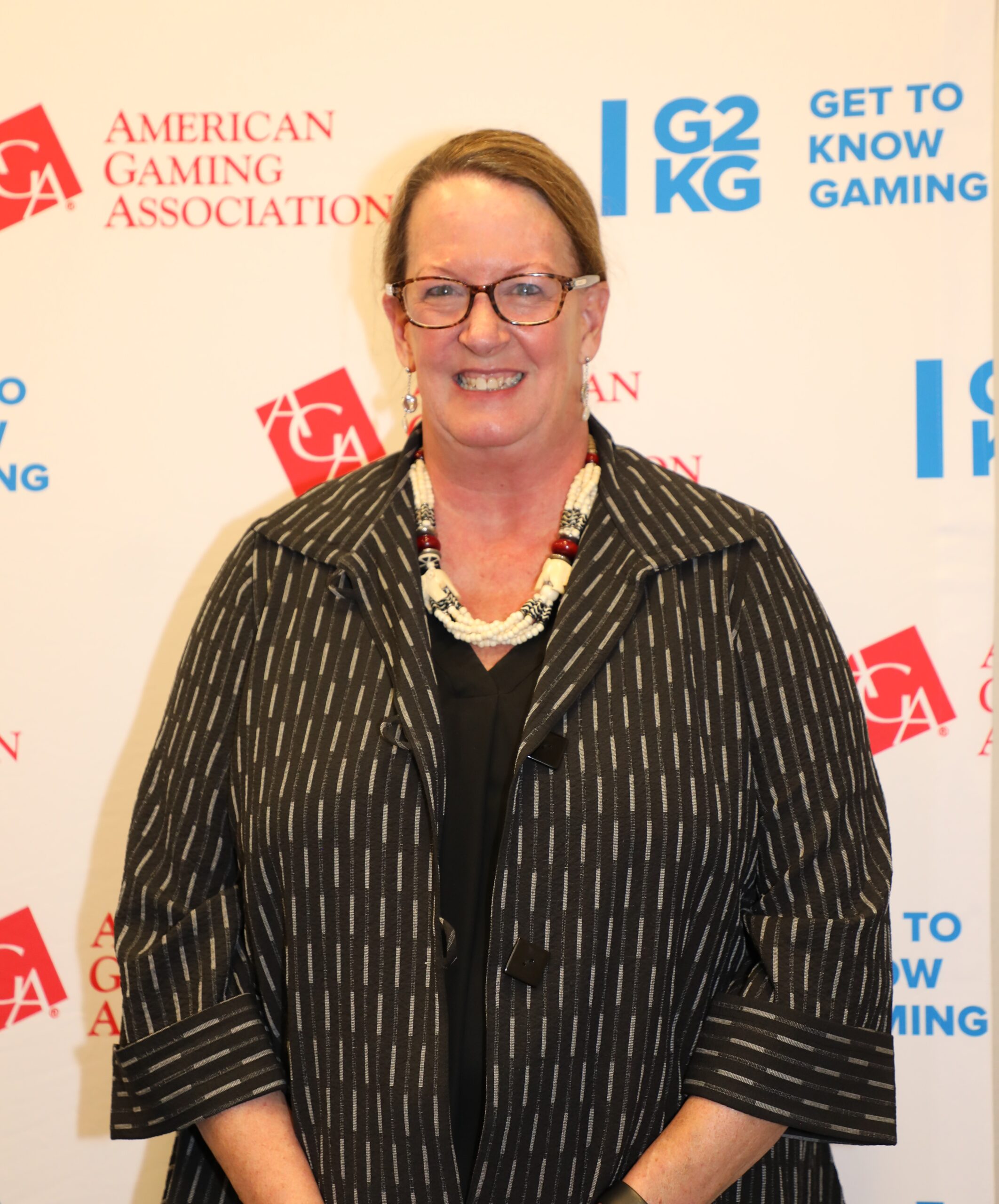 A woman wearing a striped jacket and a necklace is smiling in front of a backdrop with logos of the American Gaming Association and phrases like "Get to Know Gaming.