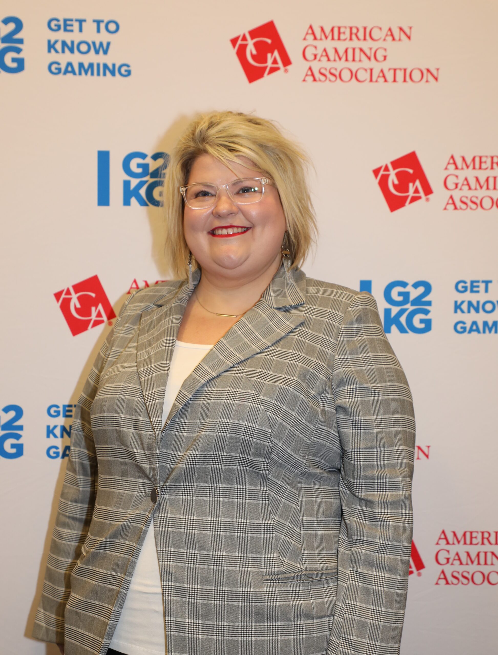 A person with short blonde hair and glasses smiles while wearing a gray plaid blazer over a white top. They are standing in front of a backdrop displaying the American Gaming Association logo and "Get to Know Gaming.