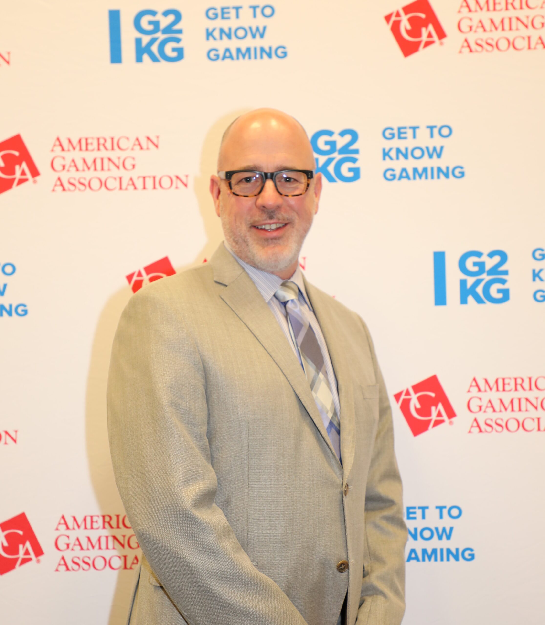 A man in a light gray suit, glasses, and a striped tie stands in front of a backdrop with logos of the American Gaming Association and "Get to Know Gaming." He is smiling and looking toward the camera.