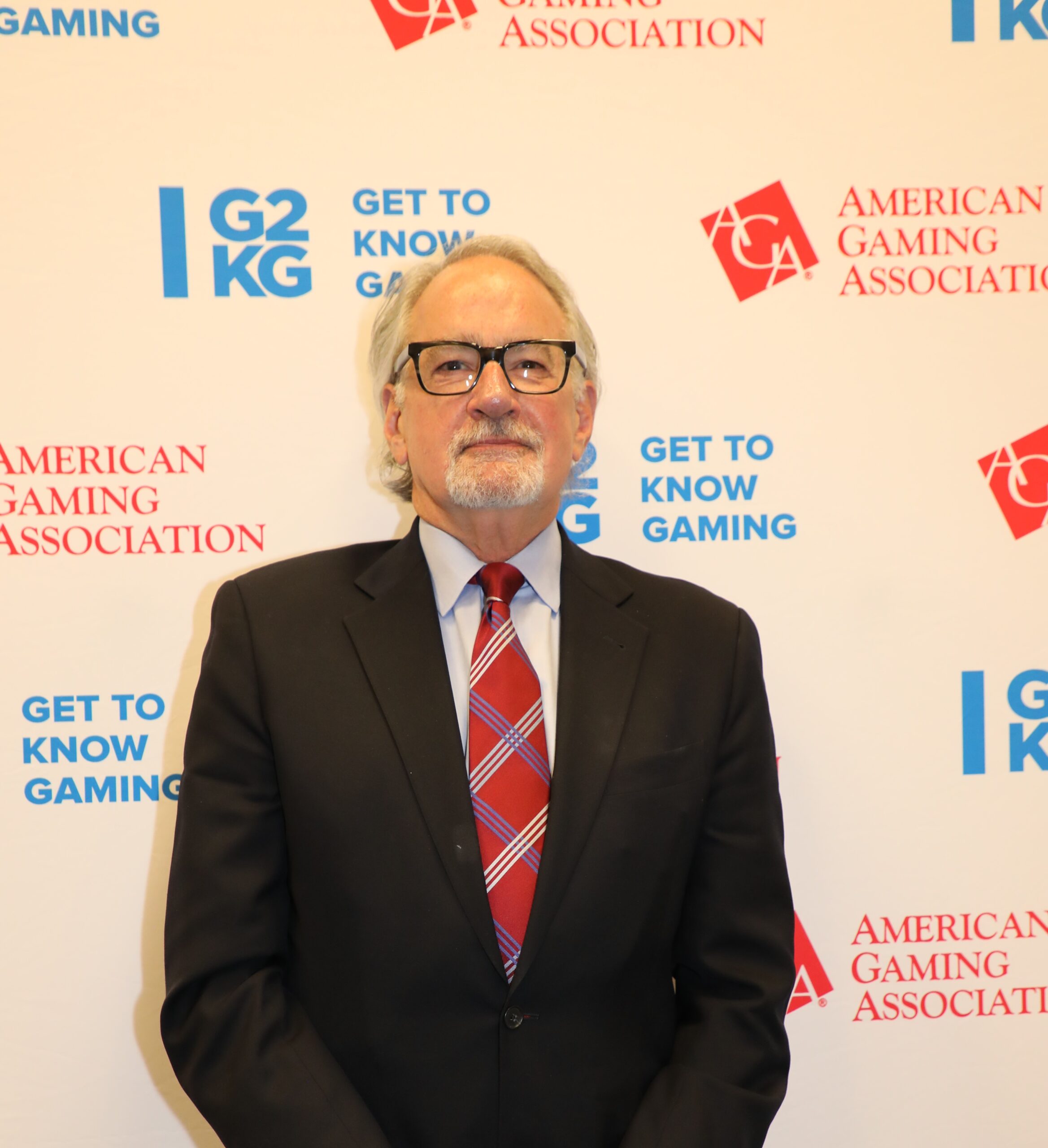 An older man with white hair and glasses stands in front of a backdrop with logos that say "American Gaming Association" and "Get to Know Gaming." He is wearing a black suit, white shirt, and red plaid tie.