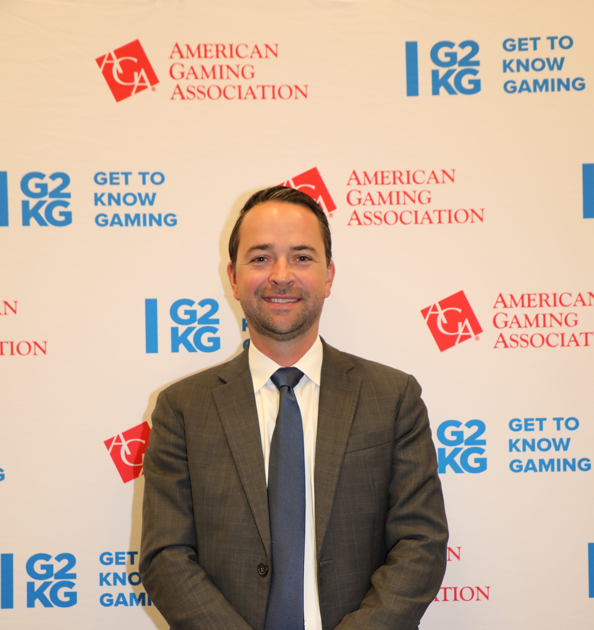 A man in a suit and tie stands in front of a backdrop with the American Gaming Association and G2KG logos repeatedly printed on it.