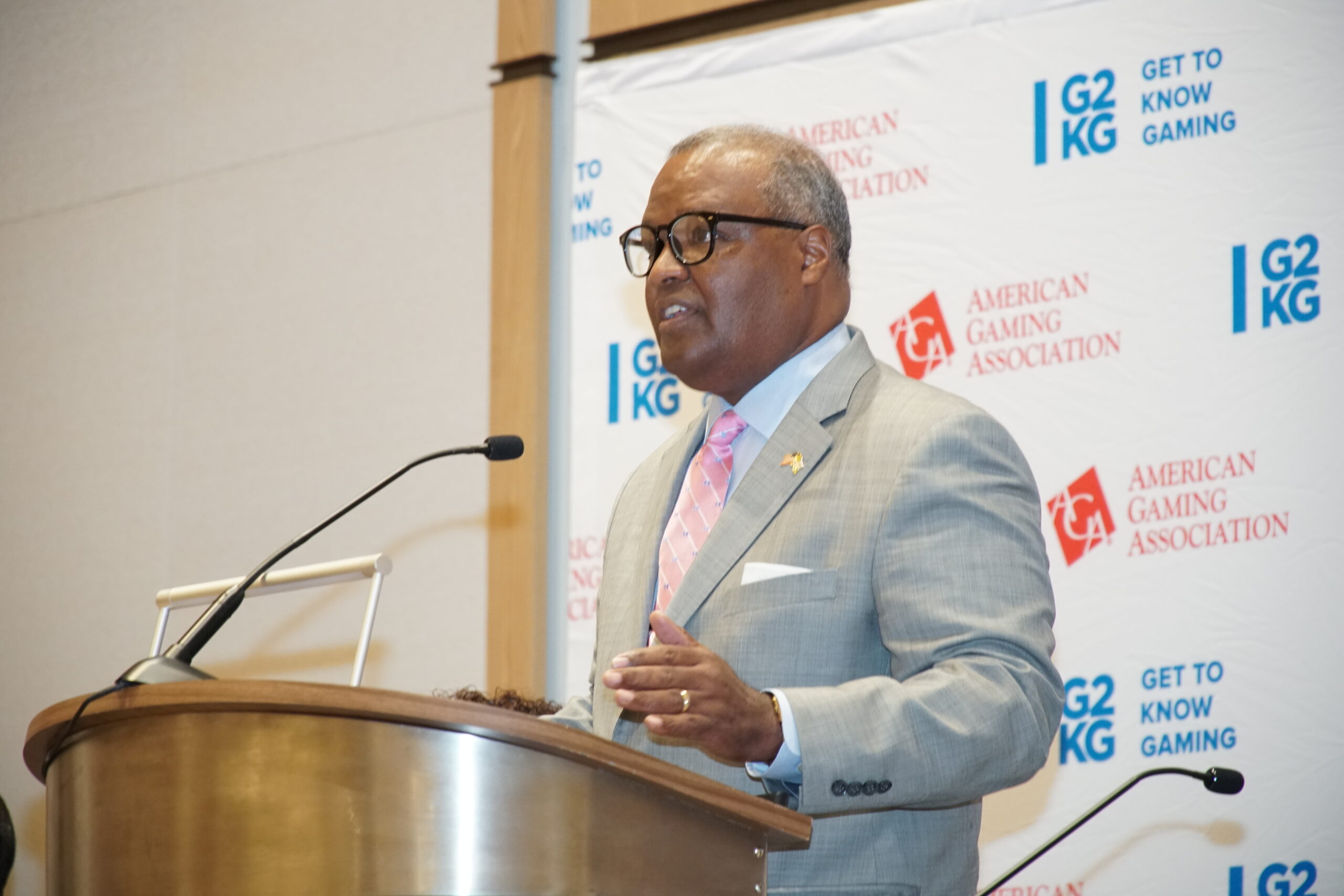 A man in a light gray suit and pink tie is speaking at a podium. Behind him is a backdrop with logos for "American Gaming Association" and "Get to Know Gaming." He's gesturing with one hand while holding a microphone with the other.
