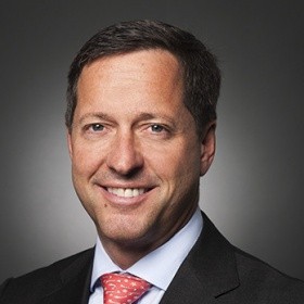 Smiling man in a suit with a light blue shirt and red tie, against a dark gray background.