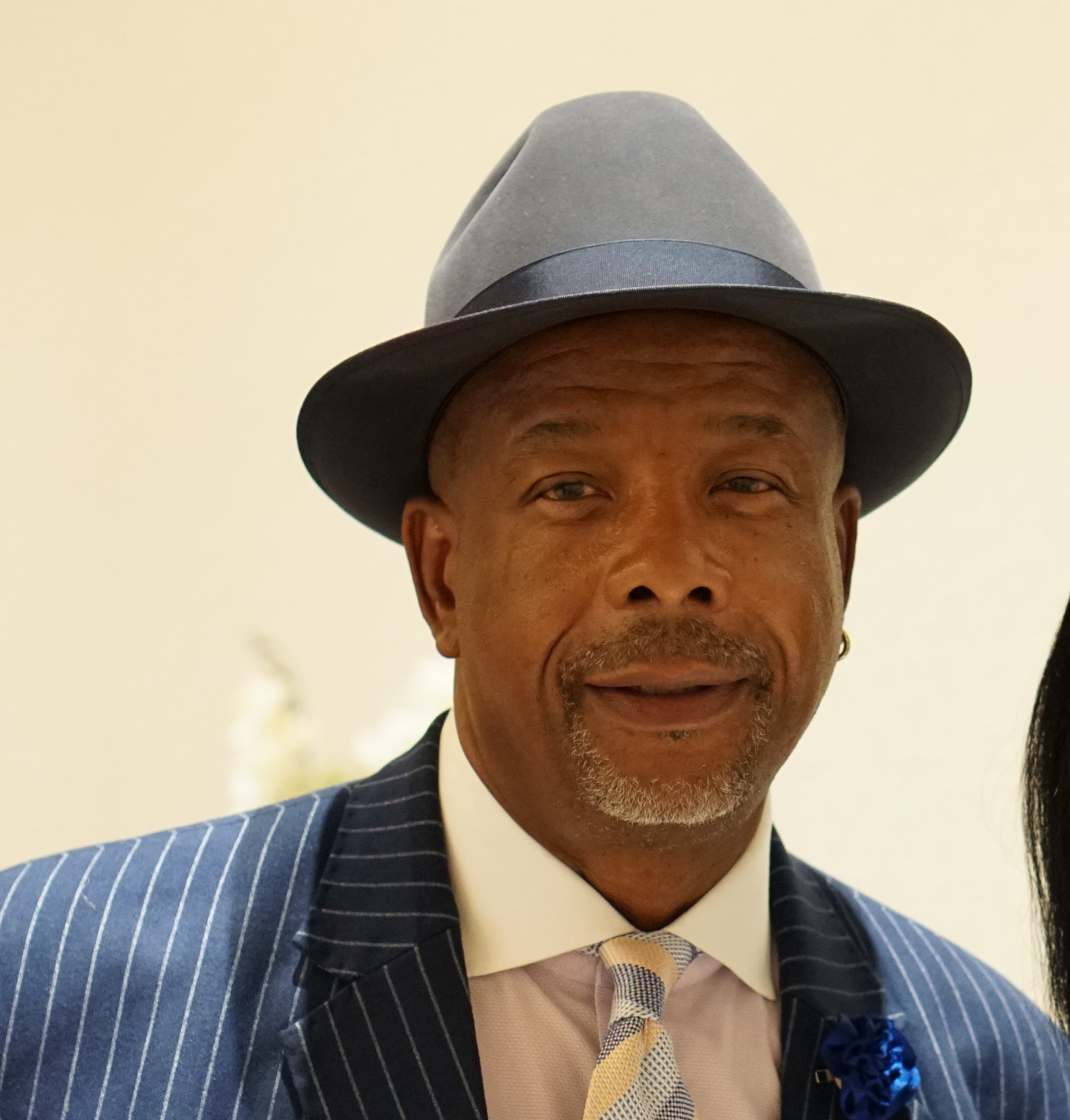 A man wearing a pinstripe suit, patterned tie, and a fedora hat is smiling. He has a small earring and a mustache. The background is neutral.