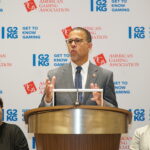 A man in a suit speaks at a podium with the "American Gaming Association" and "Get to Know Gaming" logos as a backdrop. Two women sit beside him, listening attentively.