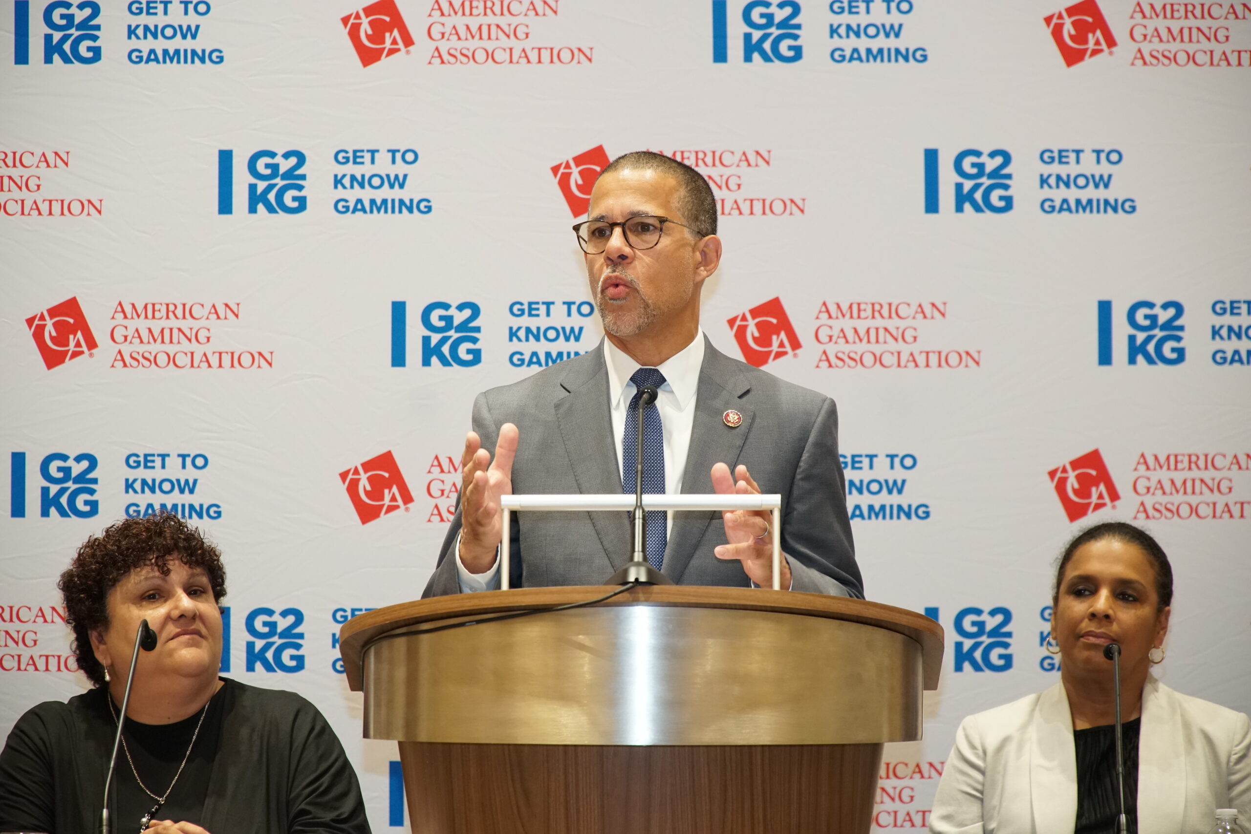 A man in a suit speaks at a podium with the American Gaming Association and Get to Know Gaming logos behind him. Two women are seated on either side, listening attentively.