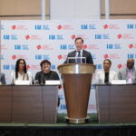 A group of people are sitting at a long table with microphones. One person is standing at a podium speaking. Behind them is a backdrop with logos, including "American Gaming Association" and "G2E.