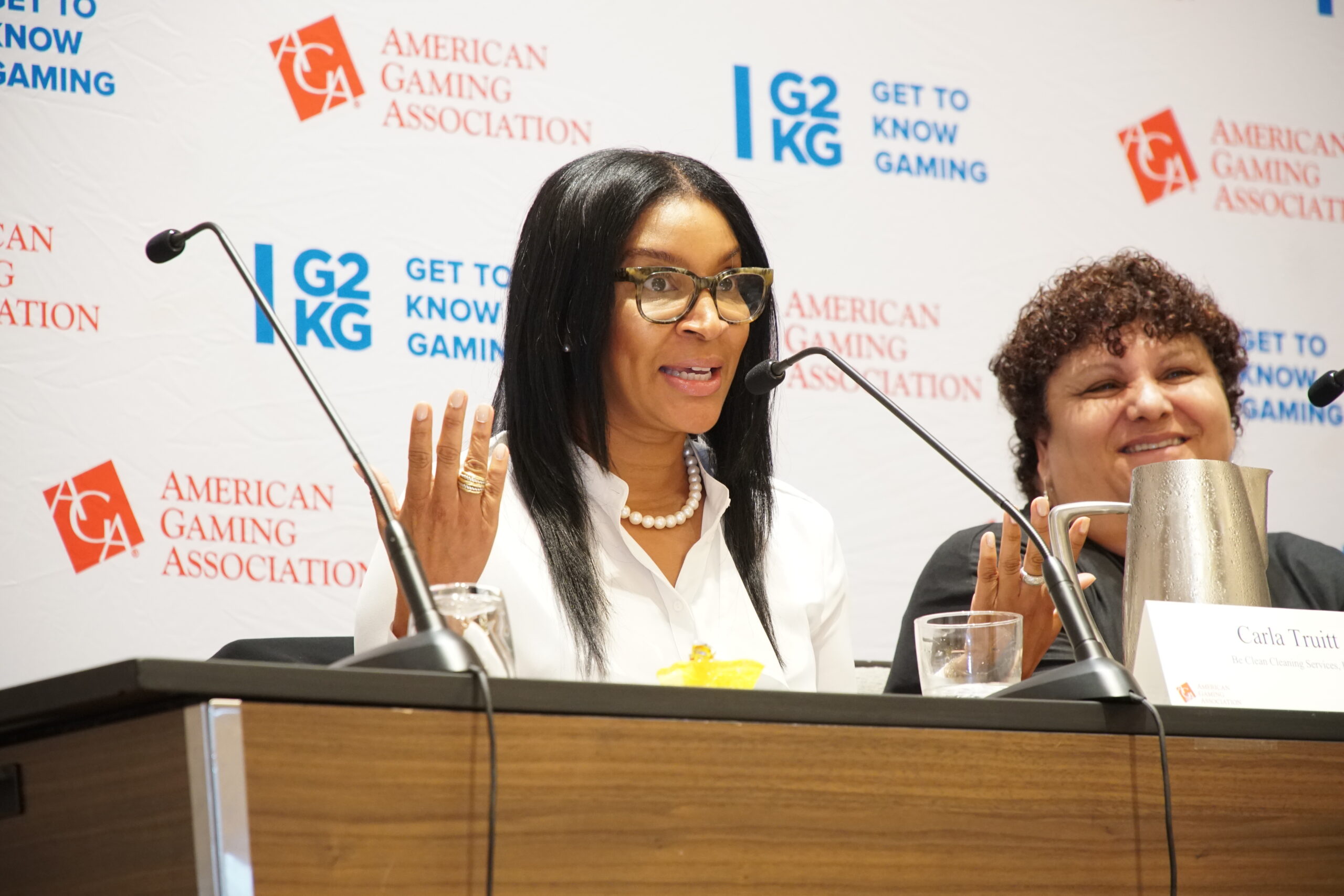 Two women sit at a panel discussion for the American Gaming Association. The woman in front speaks animatedly with hand gestures, wearing a white blouse and pearls. A microphone and nameplate are on the table. Logos and a "Get to Know Gaming" backdrop are visible.