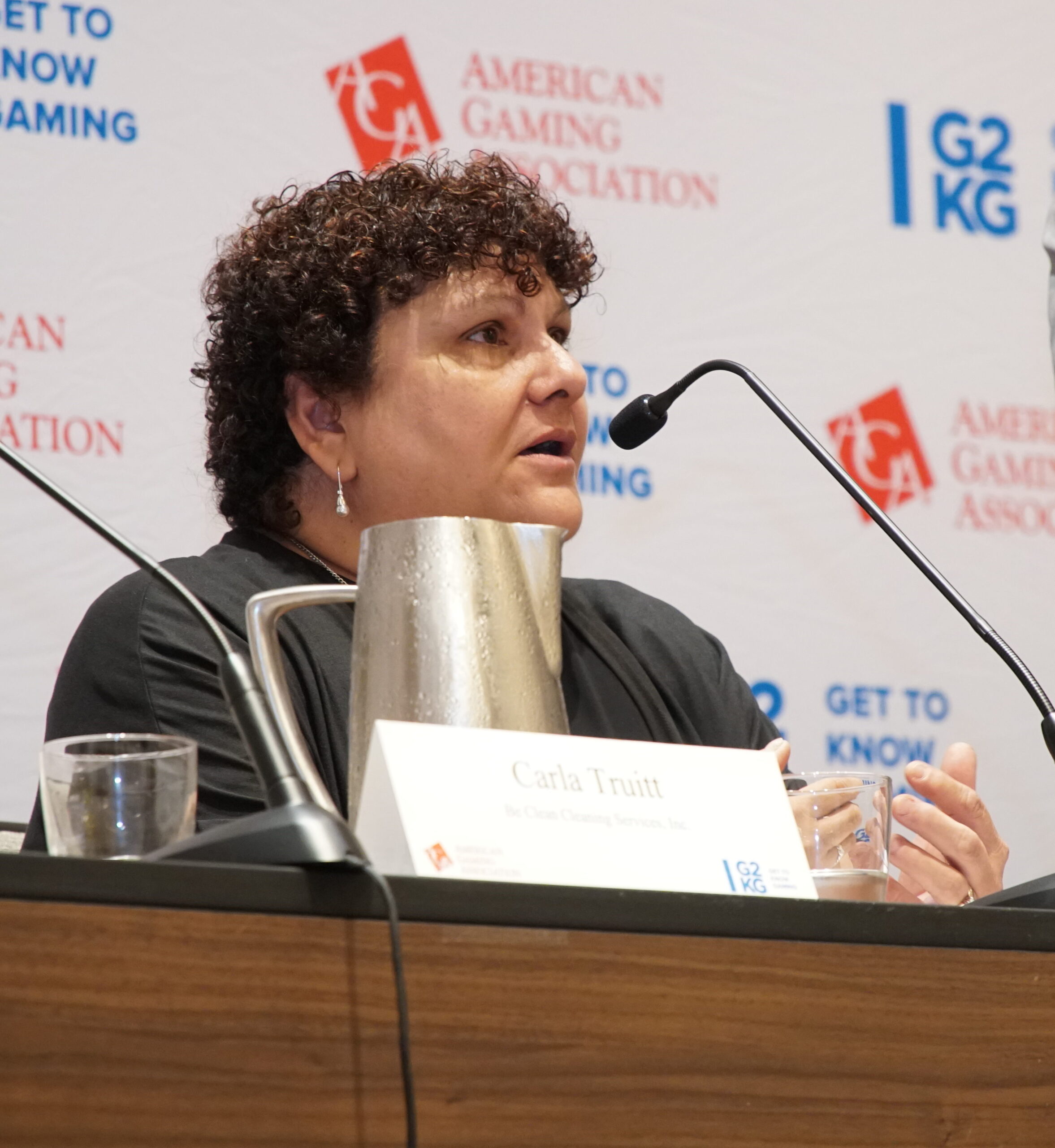 A person with curly hair speaks into a microphone at a panel. A nameplate reads "Carla Truitt" with a company name beneath it. The background has logos of the American Gaming Association and G2E. A metal water pitcher and a glass are on the table.