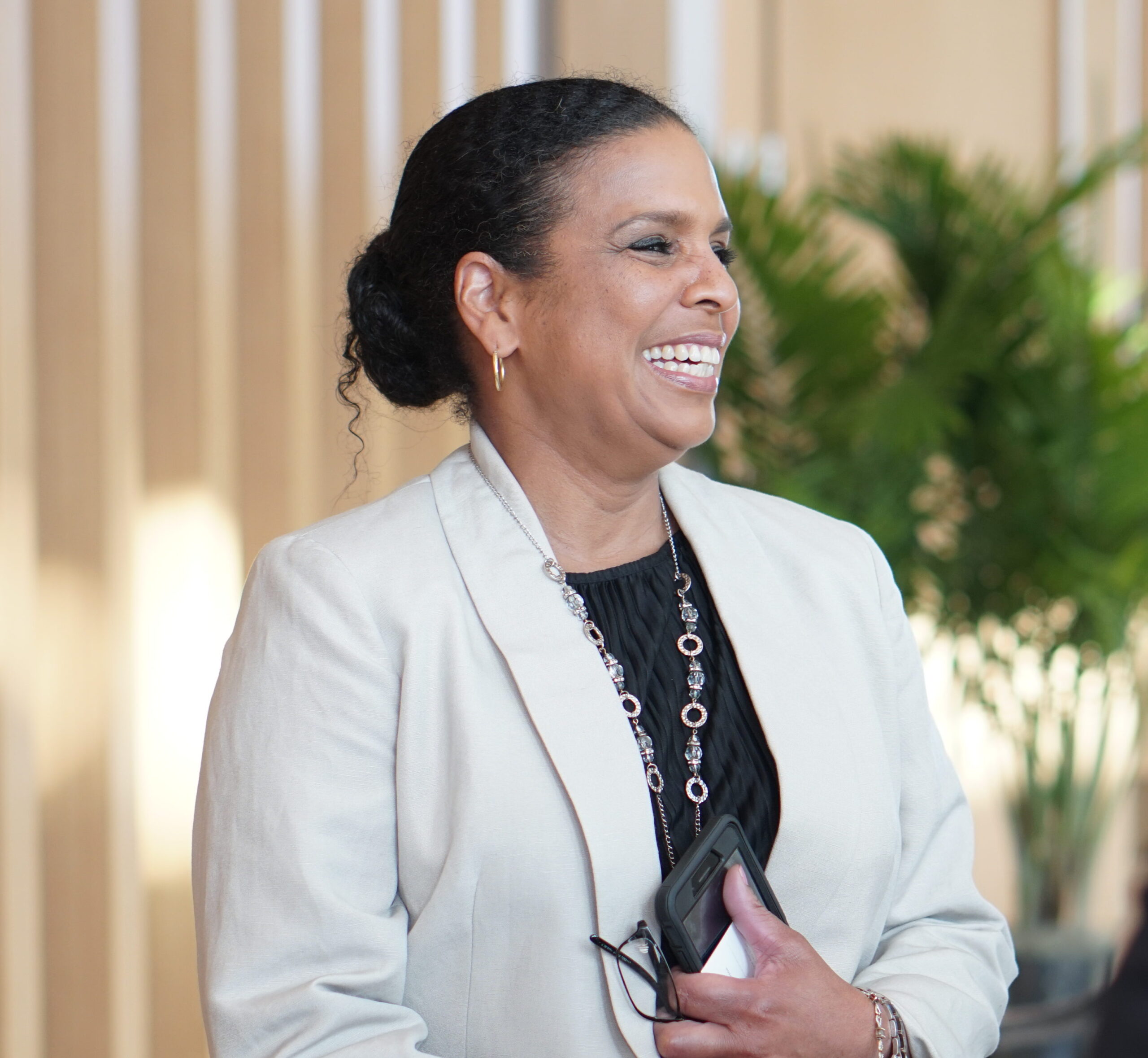 A person in a light blazer smiles while holding a phone and glasses. They are standing in a room with wooden paneling and a plant in the background.