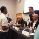 A young man interacts with panelists seated at a table during an event. The table has name plaques and water bottles. A screen in the background displays "UNLV International Gaming Institute." People are engaged and conversing.