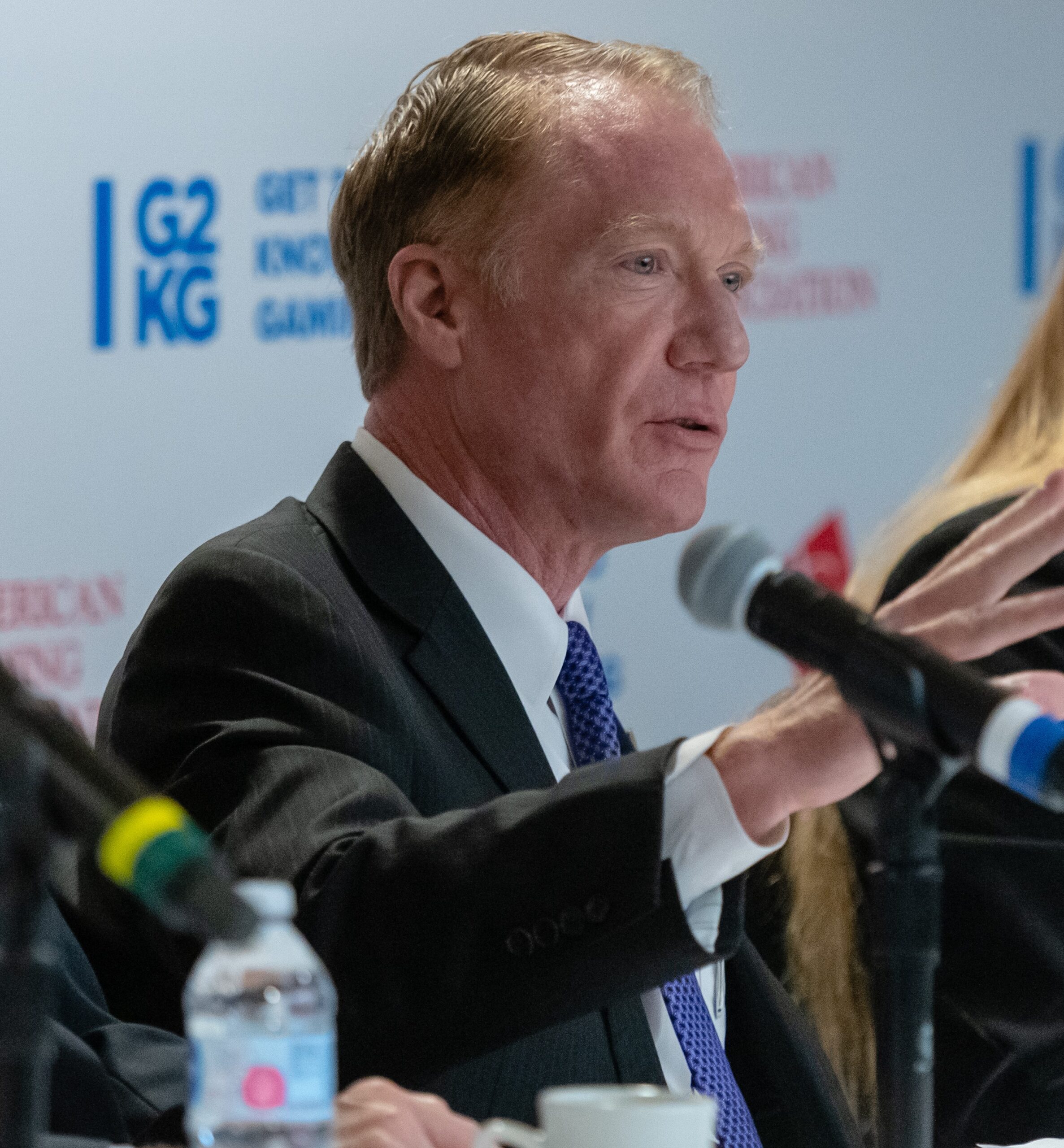 Man in a suit speaking into a microphone at a conference panel, with a small water bottle and coffee cup in front. There is a logo in the background with "G2KG" and other text partially visible.