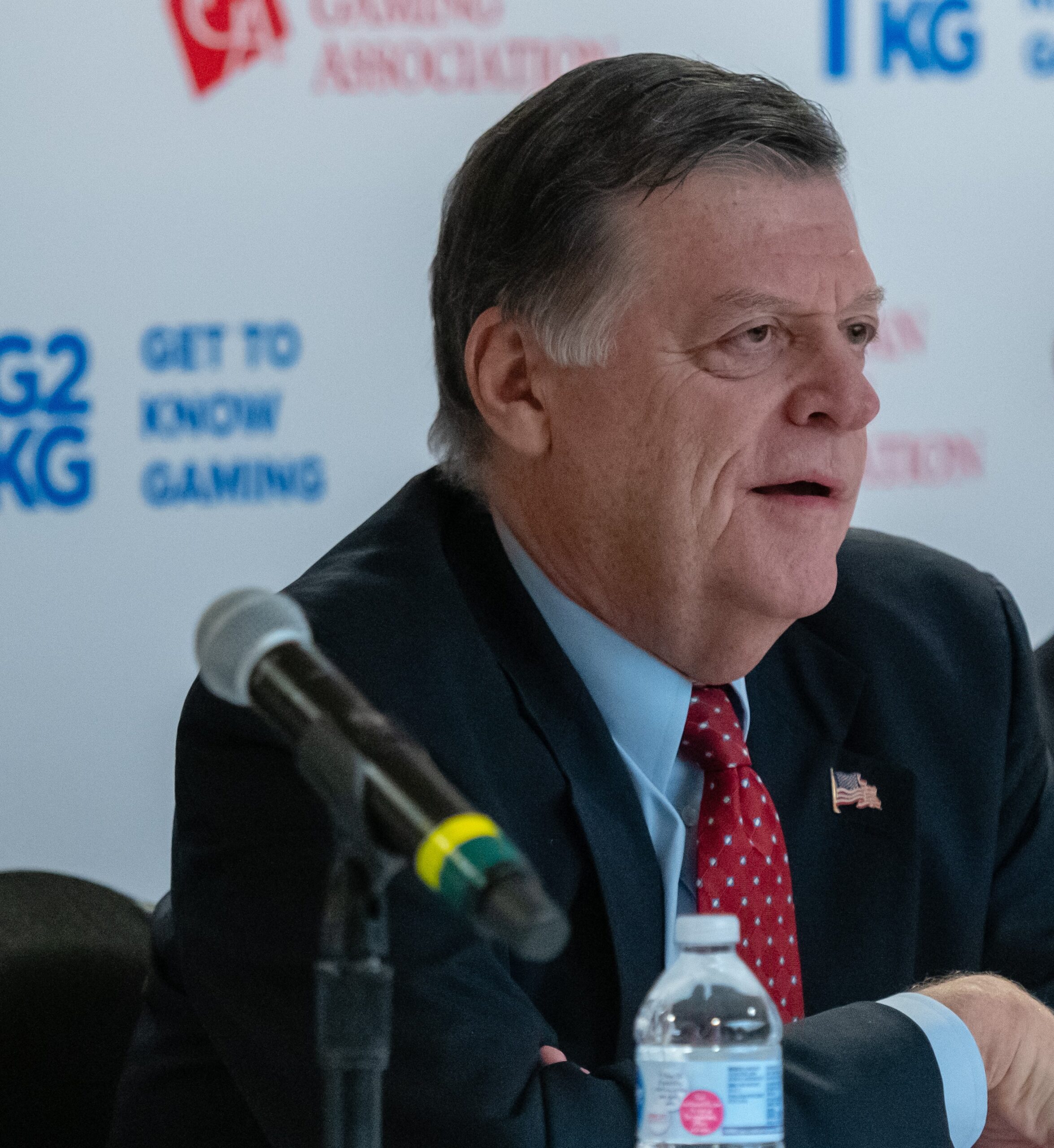 A man in a suit and red tie speaks at a panel. He is seated at a table with a microphone and a water bottle in front of him. The backdrop features logos and text, including "Get to Know Gaming.