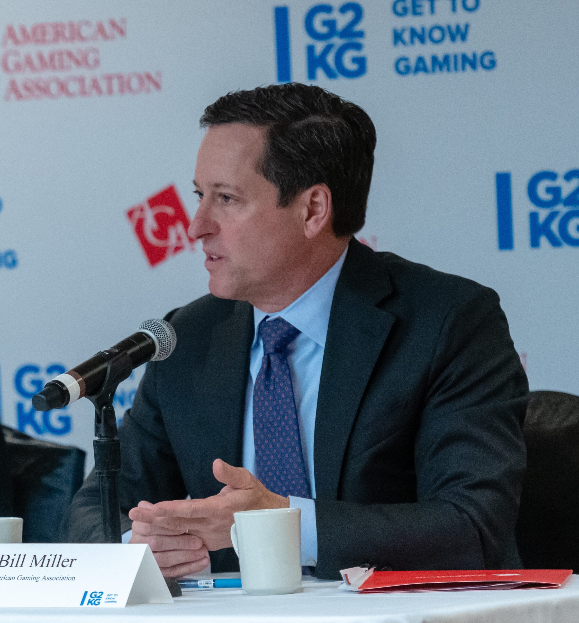Man in a suit speaking at a panel discussion with a microphone. A sign on the table says "Bill Miller," and the backdrop has logos reading "G2KG" and "American Gaming Association." A coffee cup and folder are also on the table.