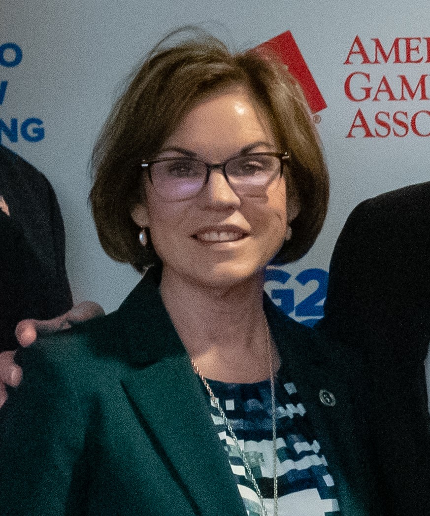A woman with short brown hair and glasses is smiling. She is wearing a dark green blazer over a striped blouse. The background features logos and text but is partially obscured.