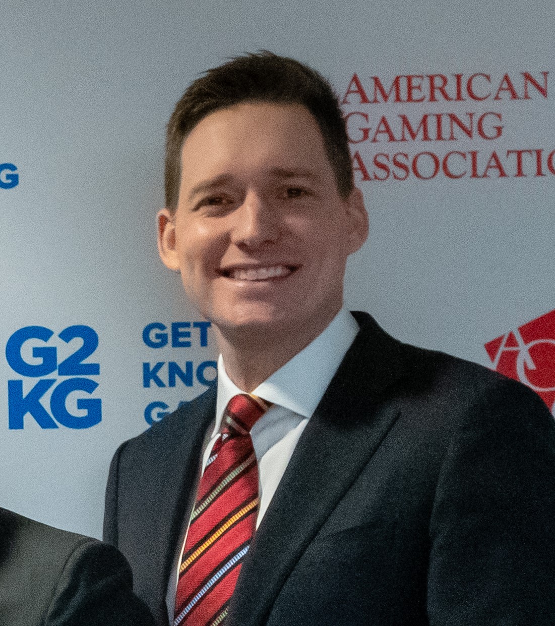A person in a suit and striped tie smiles in front of a backdrop with logos and text, including "G2KG" and "American Gaming Association.