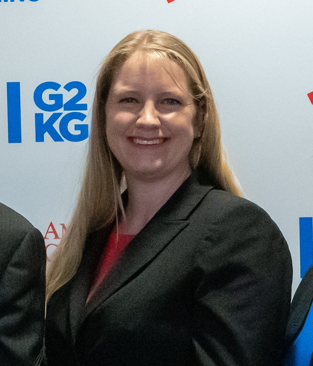 Smiling woman with long blonde hair wearing a black blazer stands in front of a backdrop with blue text "G2 KG".