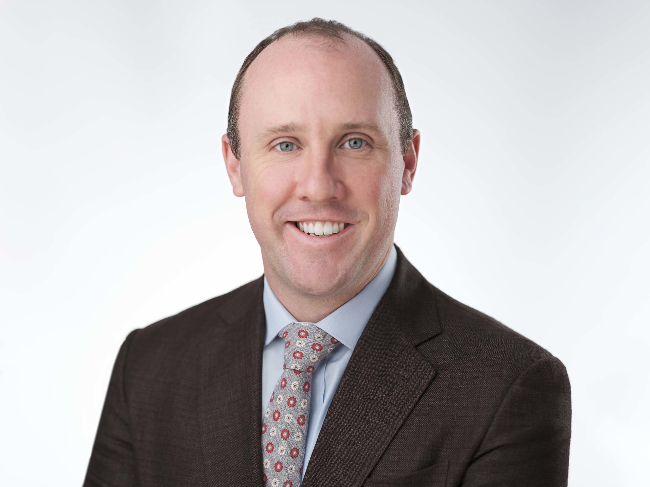 An individual wearing a brown suit, light blue shirt, and patterned tie smiles at the camera against a plain, light-colored background.