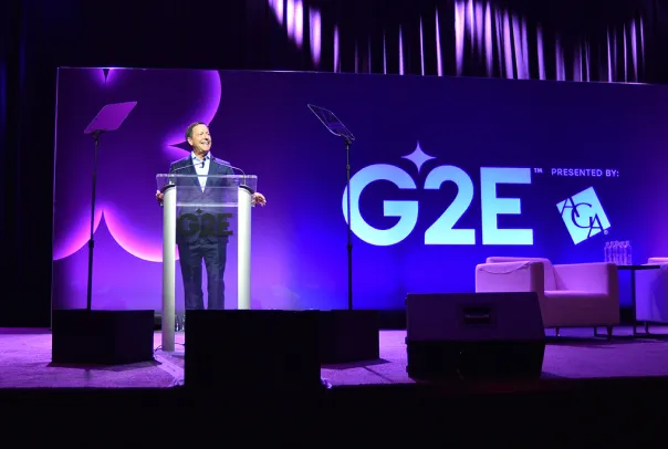 A person stands at a podium on a stage with a purple backdrop displaying "G2E" and logos. The stage is set with clear screens and a white chair. The person is speaking into a microphone.