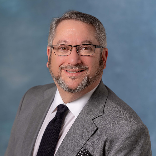 A man with short gray hair and a beard, wearing glasses, a gray suit, white shirt, and black tie, smiles at the camera. The background is a soft blue-gray gradient.