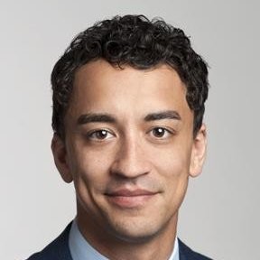 A person with short curly hair is smiling slightly at the camera. They are wearing a dark suit and a light blue shirt. The background is plain and light-colored.