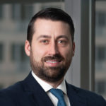 A man with dark hair and a beard is wearing a navy suit, white shirt, and blue tie, smiling in an office setting with a blurred windowed building in the background.