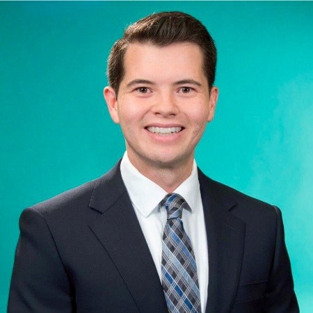 A man in a dark suit, white shirt, and plaid tie smiles against a teal background.