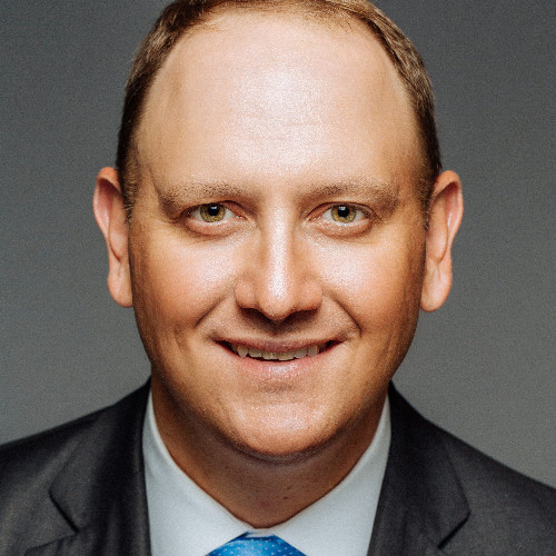 A man wearing a dark suit, white shirt, and blue tie smiles while looking directly at the camera. The background is a plain gray color.