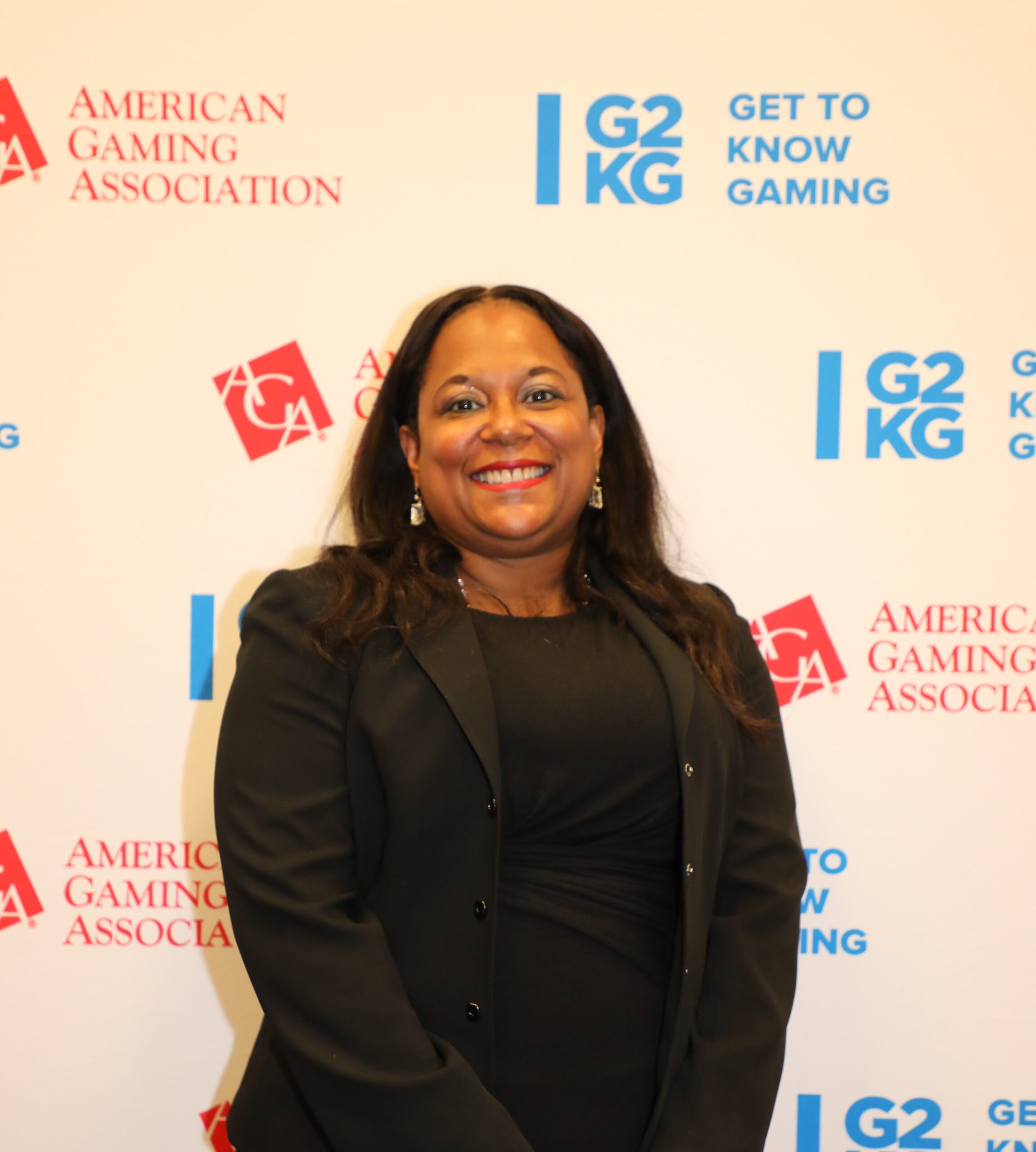 A person smiling and standing in front of a backdrop displaying logos for the American Gaming Association and Get to Know Gaming. They are wearing a black blazer and have long, dark hair.