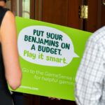 People holding a bright green sign that reads, "Put your Benjamins on a budget. Play it smart," with a website link for gaming tips.