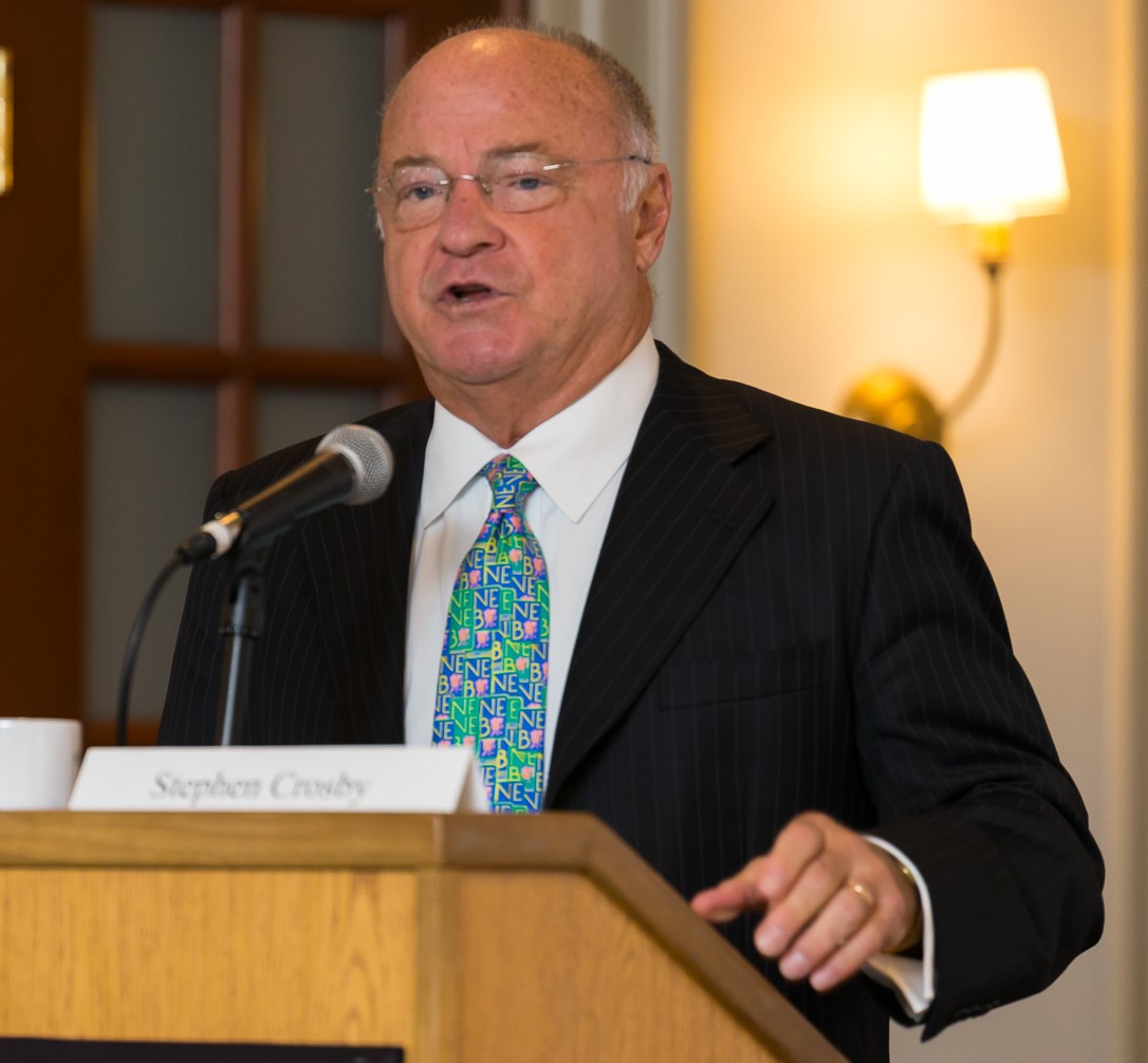An older man wearing a suit and colorful tie speaks at a podium with a microphone. A name tag on the podium reads "Stephen Crosby." There's a wall lamp and a door in the background.