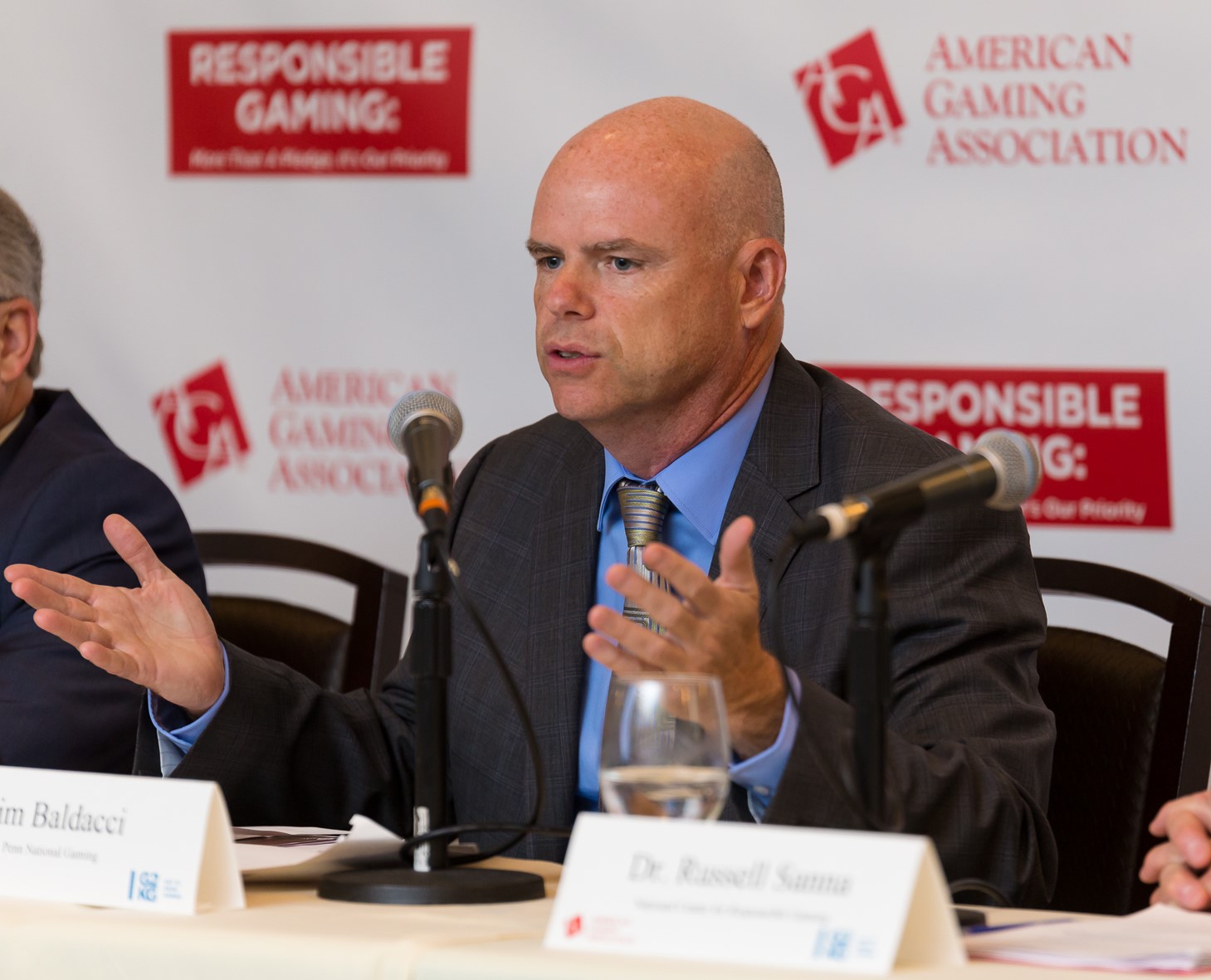 A bald man in a suit gestures while speaking at a panel discussion. Behind him, banners display "Responsible Gaming" and "American Gaming Association." A microphone and a name card are on the table in front of him.