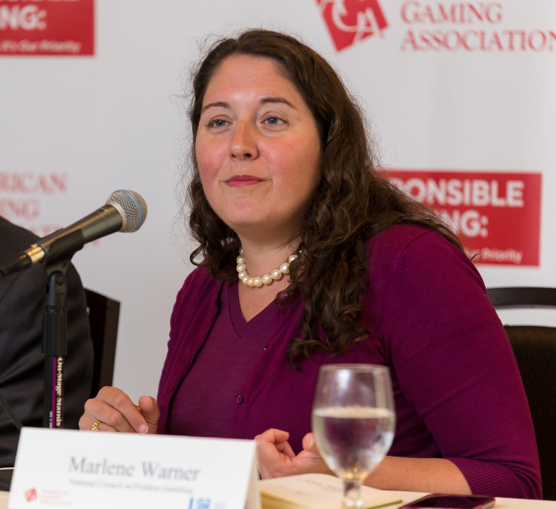 A woman with long brown hair and a maroon top speaks into a microphone at a panel event. There's a glass of water and some papers in front of her. A banner in the background reads "Gaming Association" and "Responsible Gaming".