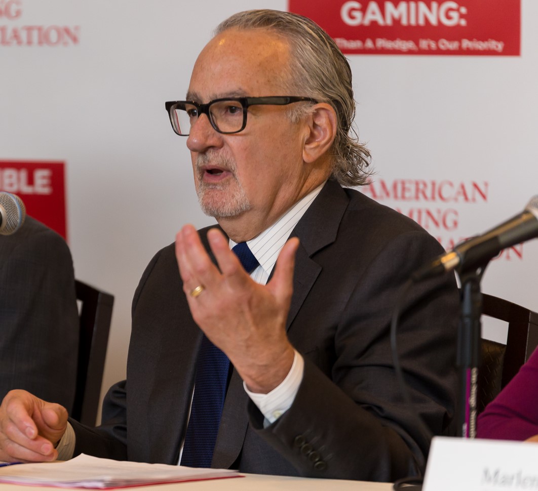 An elderly man with gray hair and glasses speaks at a conference. He is wearing a suit and tie and is gesturing with one hand. Microphones are in front of him, and a red and white conference banner is in the background.