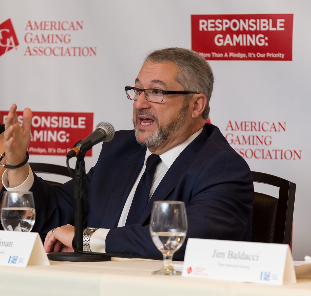 A man in a suit speaks into a microphone at a conference table with water glasses in front. Behind him are banners with "Responsible Gaming: More Than a Pledge, It's Our Priority" and "American Gaming Association" logos.