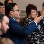 A man in a suit raises his hand while speaking in a classroom or meeting setting. Other seated individuals listen attentively, including a woman in a patterned blouse and a bearded man in the foreground. There are a few people in the background.
