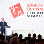 A man stands at a podium, speaking at the Sports Betting Executive Summit. The ACA logo is displayed prominently on a screen behind him. Audience members are seated in the foreground.