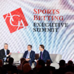 Three men in suits sit on a stage in front of a backdrop with the American Gaming Association logo and the text "Sports Betting Executive Summit." They are engaged in conversation, with one gesturing as he speaks. An audience is visible in the foreground.