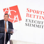 A man in a suit stands at a clear podium, speaking at the Sports Betting Executive Summit. Behind him is a logo with the letters AGA.