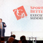 A speaker stands at a podium on a stage at the Sports Betting Executive Summit. Behind him is a large screen displaying the event's logo and name. Audience members are seated in the foreground.